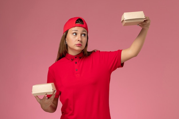 Mensageira de frente com uniforme vermelho e capa segurando pacotes de comida de entrega na parede rosa claro, trabalhador de empresa de serviço de entrega de uniforme