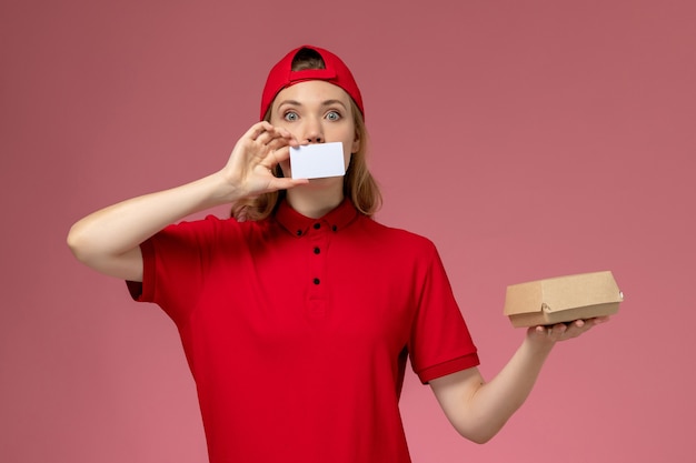 Mensageira de frente com uniforme vermelho e capa segurando um pequeno pacote de comida de entrega com um cartão de plástico branco na mesa rosa.