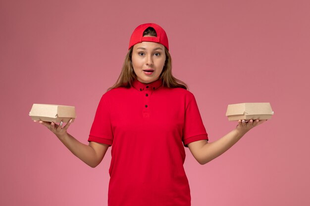 Mensageira de frente feminina em uniforme vermelho e capa segurando pacotes de entrega de comida na parede rosa claro, garota de serviço de empresa de serviço de entrega de uniforme