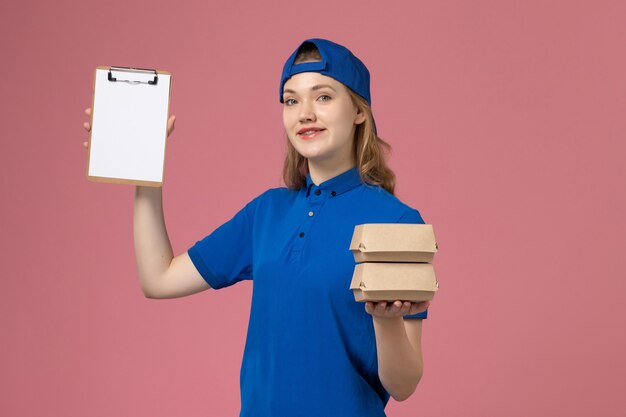 Mensageira de frente para mulher com capa uniforme azul segurando pequenos pacotes de comida para entrega e um bloco de notas sobre fundo rosa.