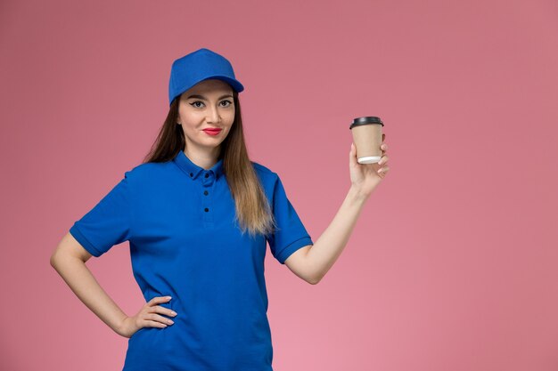 Mensageira de frente para mulher com uniforme azul e capa segurando a xícara de café de entrega posando na parede rosa