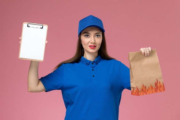 Mensageira de frente para mulher com uniforme azul e capa segurando o bloco de notas e o pacote de comida de papel na parede rosa trabalhador
