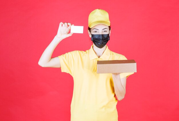 Mensageira de uniforme amarelo e máscara preta segurando a caixa de papelão e apresentando seu cartão de visita