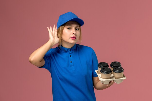 mensageira de uniforme azul segurando xícaras marrons de café tentando ouvir no rosa, trabalhador de entrega de uniforme de serviço