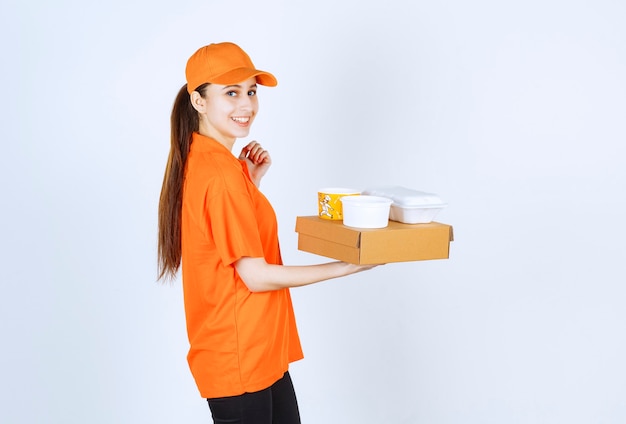 Mensageira de uniforme laranja segurando uma caixa de papelão, uma caixa de plástico para viagem e um copo de macarrão amarelo