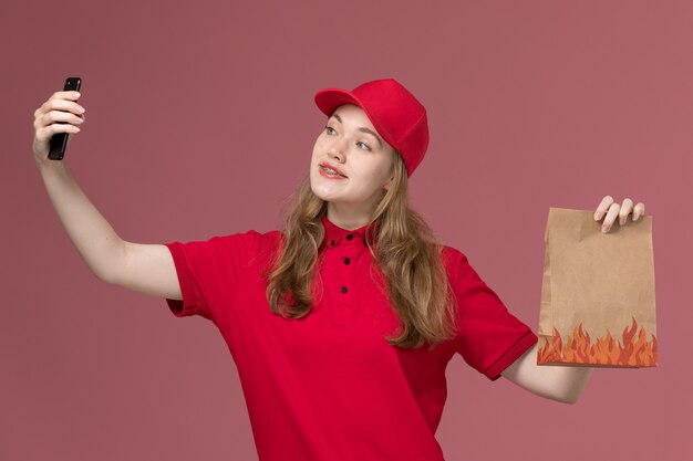 mensageira de uniforme vermelho segurando um pacote de comida e tomando selfie em rosa claro, entrega de trabalhador de serviço de uniforme de trabalho