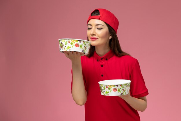 Mensageira de vista frontal com capa de uniforme vermelha e tigelas de entrega nas mãos, cheirando a parede rosa claro, funcionária do prestador de serviços