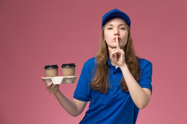 Mensageira feminina de uniforme azul segurando xícaras de café marrons, mostrando o sinal de silêncio no uniforme rosa de serviço da mesa, entregando trabalho na empresa, vista frontal