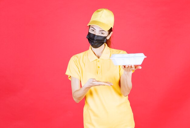 Mensageira feminina em uniforme amarelo e máscara preta segurando um pacote para viagem