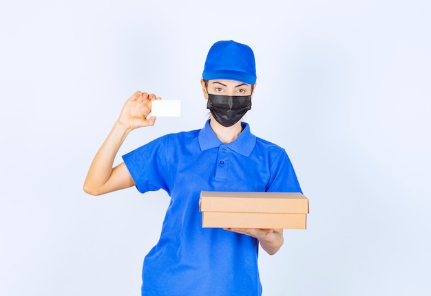 Mensageira feminina em uniforme azul e máscara facial entregando um pacote de papelão e apresentando seu cartão de visita.