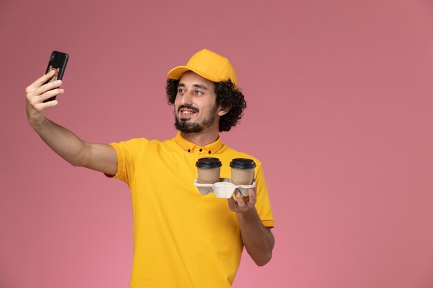 Mensageiro de frente com uniforme amarelo e capa segurando xícaras de café marrom tirando foto na parede rosa