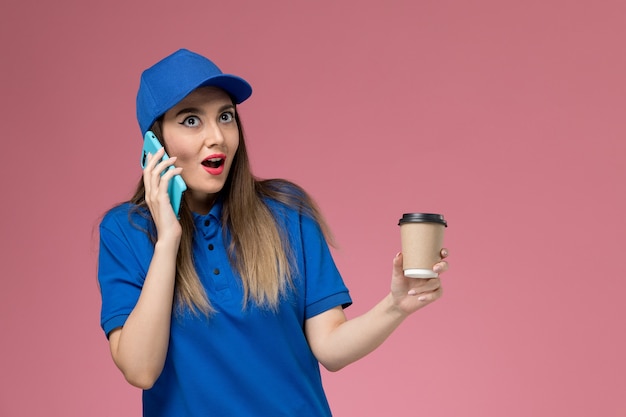 Mensageiro de frente com uniforme azul e capa segurando a xícara de café de entrega usando um telefone na parede rosa