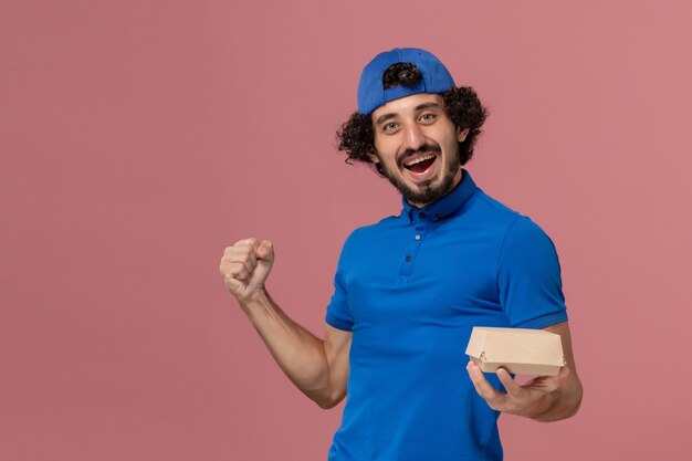 Mensageiro de frente com uniforme azul e capa segurando um pequeno pacote de comida para entrega e regozijando-se com a empresa de serviço de entrega de uniforme de parede rosa