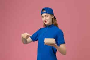 Foto grátis mensageiro de frente com uniforme azul e capa segurando um pequeno pacote de entrega de comida no fundo rosa uniforme de entrega serviço empresa trabalhador