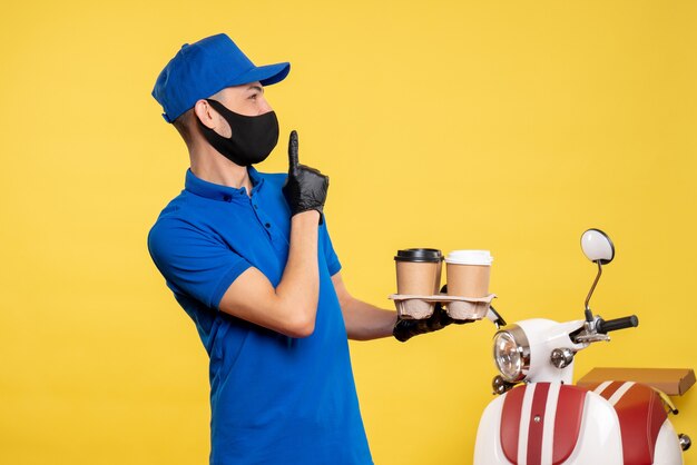 Foto grátis mensageiro de frente com uniforme azul e máscara segurando café em uniforme de trabalho amarelo serviço covide - pandemia de entrega