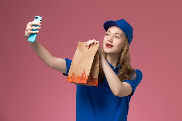 Mensageiro de frente com uniforme azul tirando uma selfie com pacote de comida na mesa de luz rosa serviço da empresa uniforme trabalho