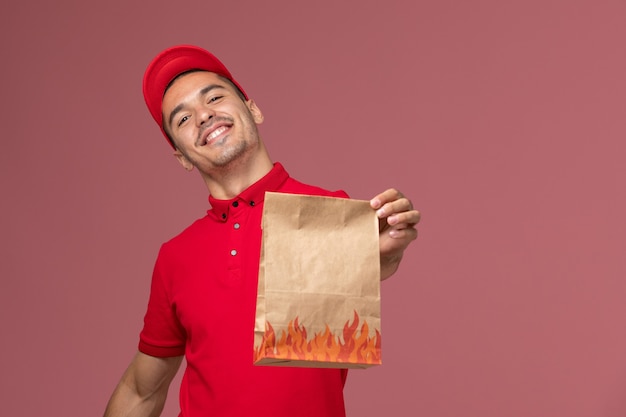 Mensageiro de frente com uniforme vermelho e capa segurando um pacote de comida de papel com um sorriso na parede rosa Serviço de entrega de uniforme masculino trabalhador