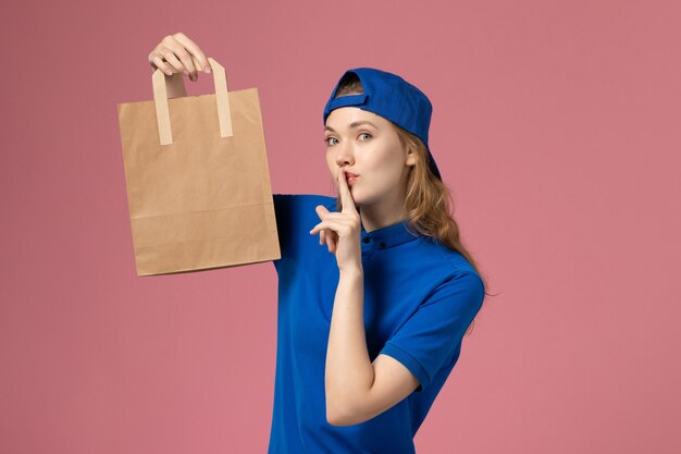 Mensageiro feminino com capa de uniforme azul segurando um pacote de papel na parede rosa, trabalho de entrega de serviço de funcionário