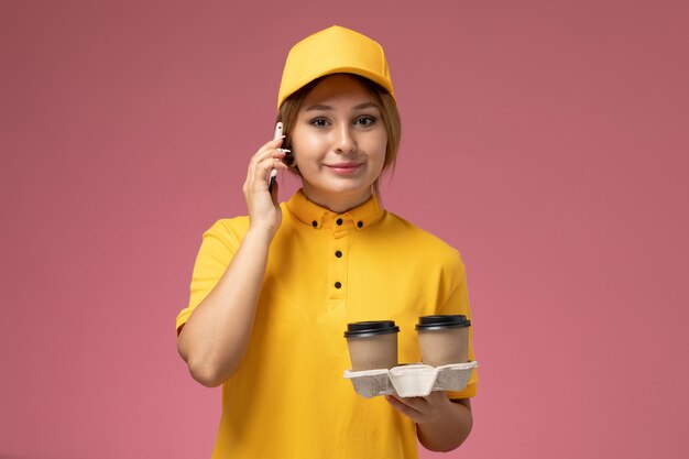 Mensageiro feminino de vista frontal em uniforme amarelo capa amarela segurando xícaras de café de plástico falando ao telefone na mesa rosa uniforme entrega trabalho cor trabalho