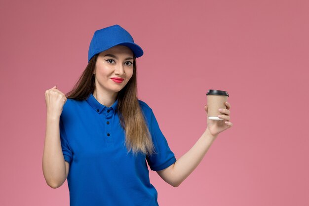 Mensageiro feminino de vista frontal em uniforme azul e capa segurando a xícara de café de entrega com expressão encantada na mulher de trabalho de mesa rosa