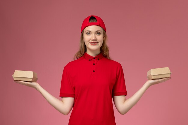 Mensageiro feminino de vista frontal em uniforme vermelho e capa segurando pequenos pacotes de comida de entrega na parede rosa, trabalhador de uniforme de empresa de serviço de entrega de trabalho