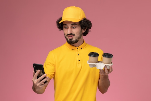 Mensageiro masculino com capa e uniforme amarelo, segurando xícaras de café marrons e usando um telefone na parede rosa