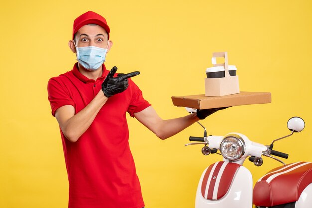 Mensageiro masculino com máscara e entrega de café e caixa em uniforme de trabalho de vírus covidêmico de serviço de cor amarela