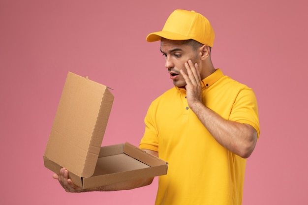 Foto grátis mensageiro masculino com uniforme amarelo segurando e abrindo a caixa de entrega de comida no fundo rosa claro