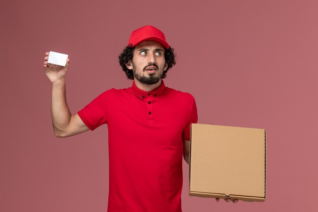 Mensageiro masculino de camisa vermelha e capa segurando a caixa de entrega de comida e o cartão na parede rosa