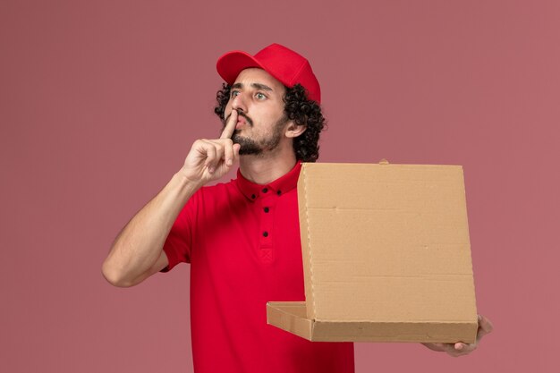 Mensageiro masculino de camisa vermelha e capa segurando a caixa de entrega de comida na parede rosa claro de frente