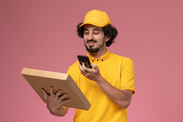 Mensageiro masculino de uniforme amarelo segurando uma caixa de entrega de comida tirando uma foto na parede rosa