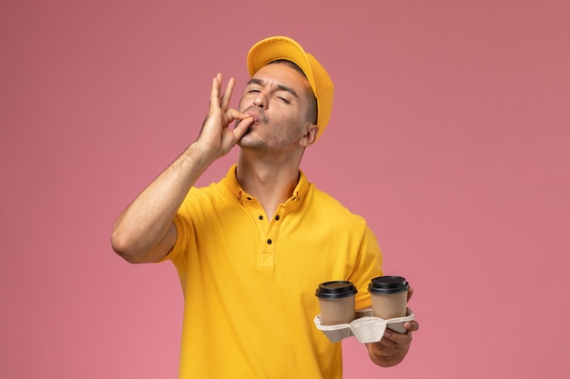 Mensageiro masculino de uniforme amarelo segurando xícaras de café de entrega mostrando uma placa saborosa em fundo rosa claro.