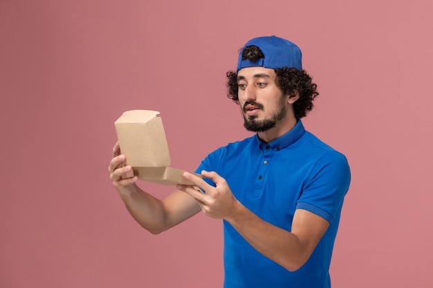 Mensageiro masculino de uniforme azul e capa segurando um pequeno pacote de entrega de comida abrindo-o na parede rosa