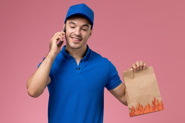 mensageiro masculino de uniforme azul segurando um pacote de comida de entrega falando no telefone na rosa