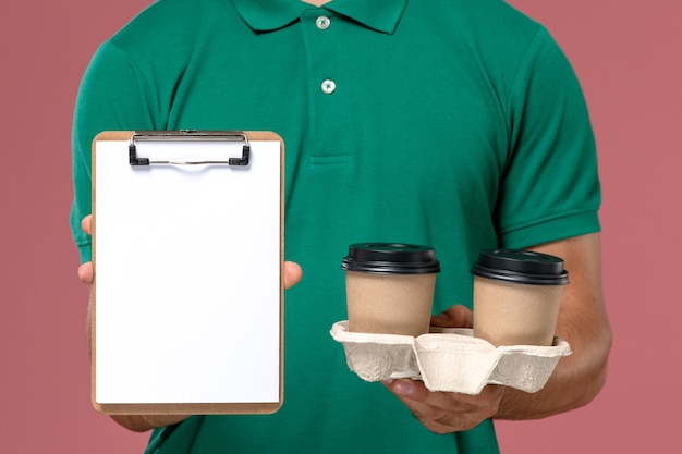 Mensageiro masculino de uniforme verde segurando xícaras de café marrom e um bloco de notas na mesa rosa