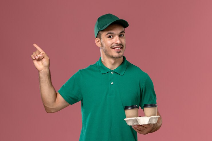 Foto grátis mensageiro masculino de uniforme verde segurando xícaras de café marrons com um sorriso na mesa rosa