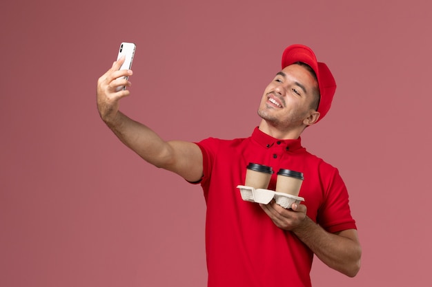Mensageiro masculino de uniforme vermelho segurando xícaras de café marrons e tirando uma selfie com eles na parede rosa