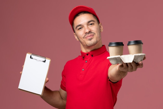 Mensageiro masculino de uniforme vermelho segurando xícaras de café marrons e um bloco de notas na parede rosa claro masculino