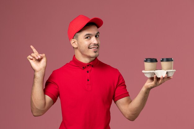 Mensageiro masculino de uniforme vermelho segurando xícaras de café marrons na mesa rosa trabalhador