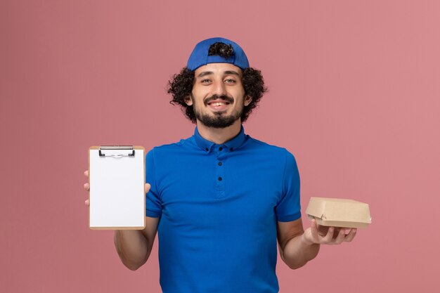 Foto grátis mensageiro masculino de vista frontal com uniforme azul e capa segurando um pequeno pacote de comida para entrega e um bloco de notas no serviço de entrega de trabalho uniforme de parede rosa