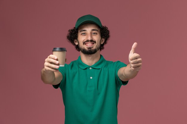 Mensageiro masculino de vista frontal com uniforme verde e capa segurando a xícara de café de entrega no trabalho de serviço de entrega uniforme de fundo rosa