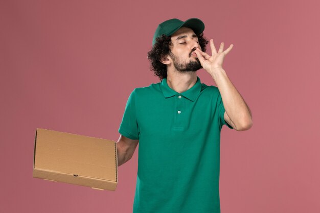 Mensageiro masculino de vista frontal com uniforme verde e capa segurando caixa de comida no fundo rosa serviço de entrega uniforme de trabalho