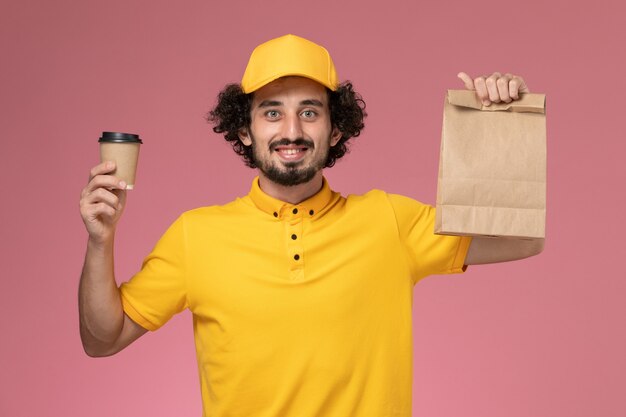 Mensageiro masculino de vista frontal em uniforme amarelo e capa segurando a xícara de café de entrega e pacote de comida na mesa rosa uniforme trabalho serviço empresa trabalhador