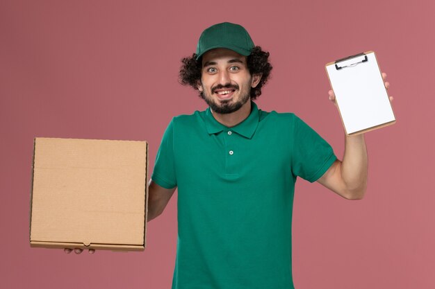 Mensageiro masculino de vista frontal em uniforme verde e capa segurando o bloco de notas de caixa de comida de entrega no fundo rosa entrega uniforme de serviço