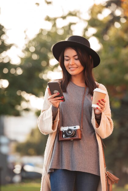 Mensagem de texto encantadora da mulher asiática do turista no telefone móvel, mantendo o parque da xícara de café em público