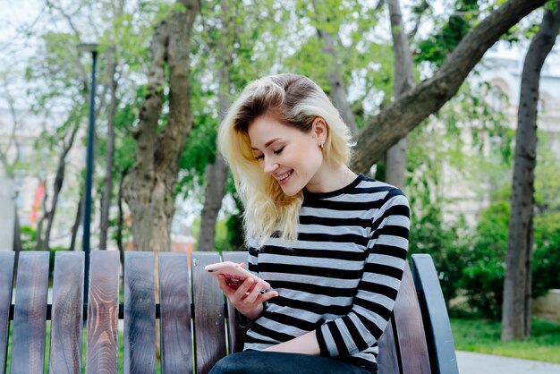 Mensagens de mulher feliz em um smartphone usando blusa listrada e sentado ao ar livre em um banco