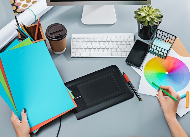 mesa cinza com laptop, bloco de notas com uma folha em branco, vaso de flores, caneta e tablet para retoque