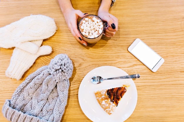 Mesa com luvas brancas de inverno, telefone, chocolate quente segurando pela garota e um pedaço de torta, chapéu cinza de malha.