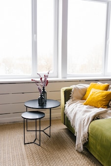 Mesa de centro preta com vasos e galhos com folhas no carpete em uma foto real do interior de uma sala de estar bem iluminada com um cobertor e almofadas amarelas em um sofá verde em uma sala de estar bem iluminada