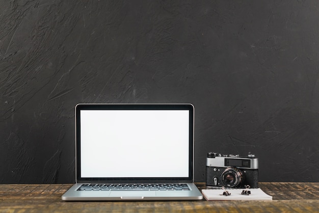 Mesa de madeira com laptop de tela em branco e câmera fotográfica retrô em fundo preto